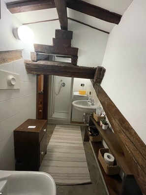 Een compacte badkamer met een plafond van houten balken, een wit wastafeltje en een klein opbergkastje.