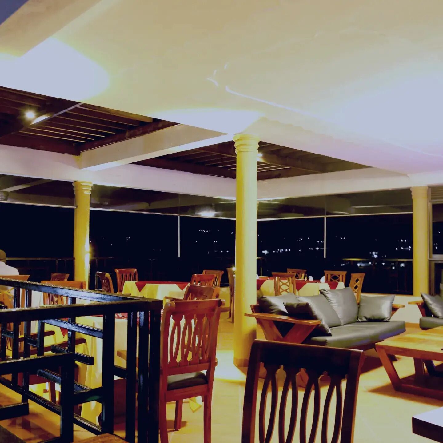 Un área de comedor al aire libre con mesas y sillas de madera, pilares amarillos y vista al cielo nocturno.