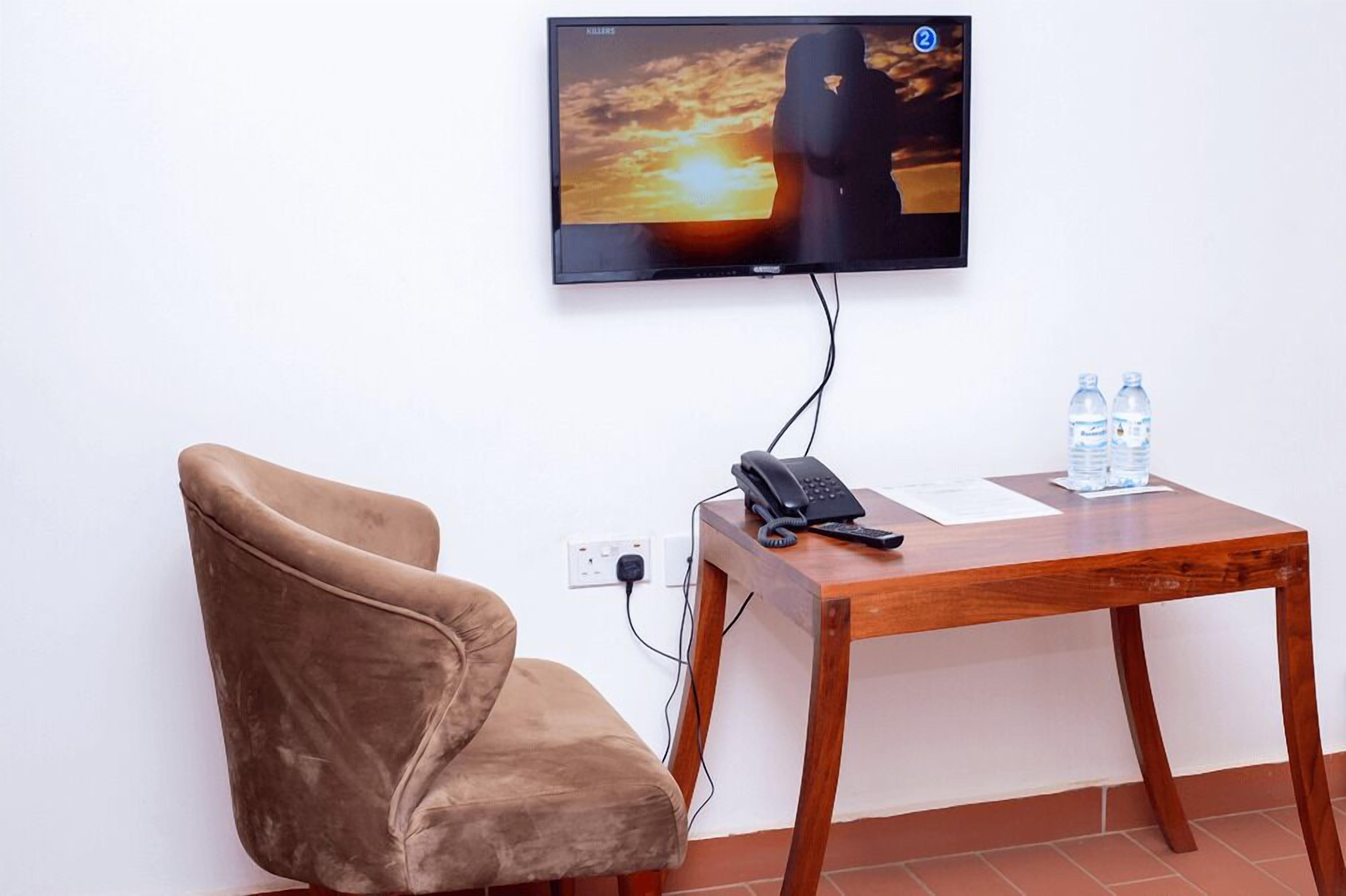 Una habitación con un televisor de pantalla plana montado en la pared mostrando una escena de atardecer, una silla marrón, una mesa de madera con un teléfono y botellas de agua.