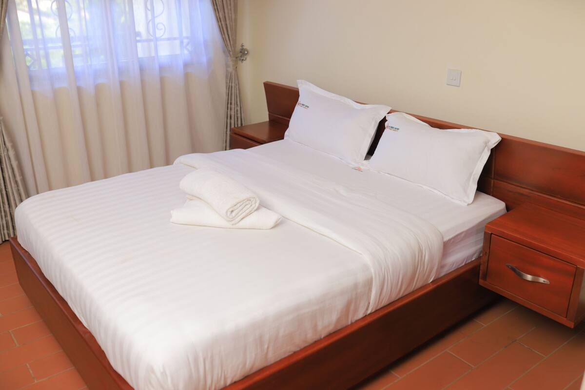 A neatly made bed with white linens and pillows, a wooden headboard, and a wooden nightstand with a drawer.