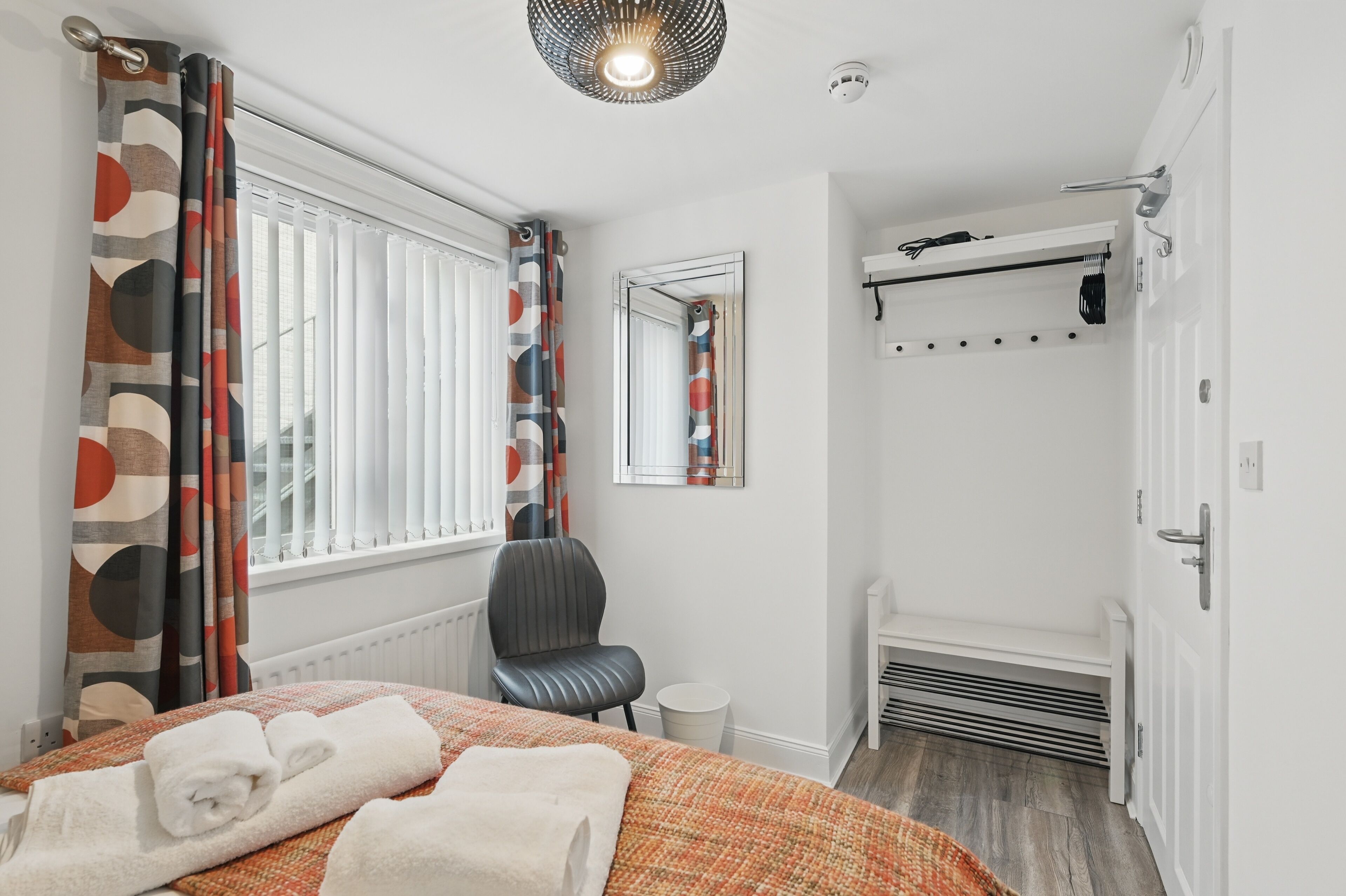 Ein Schlafzimmer mit Bett, Sessel, Heizkörper, Spiegel und einem Fenster mit Vorhängen.