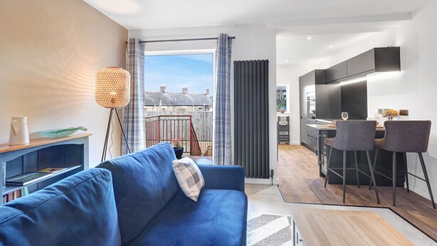 A modern apartment with a blue sofa, a dining area with two chairs, a kitchen with a TV, and a balcony with a view of the city.