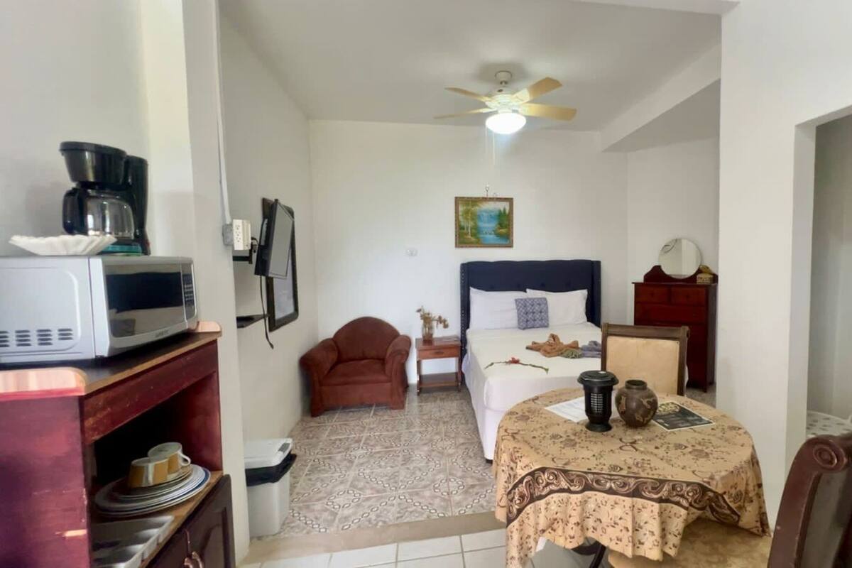 A compact hotel room with a bed, a small table with a tablecloth, a chair, a microwave, a coffee maker, a ceiling fan, and a refrigerator.