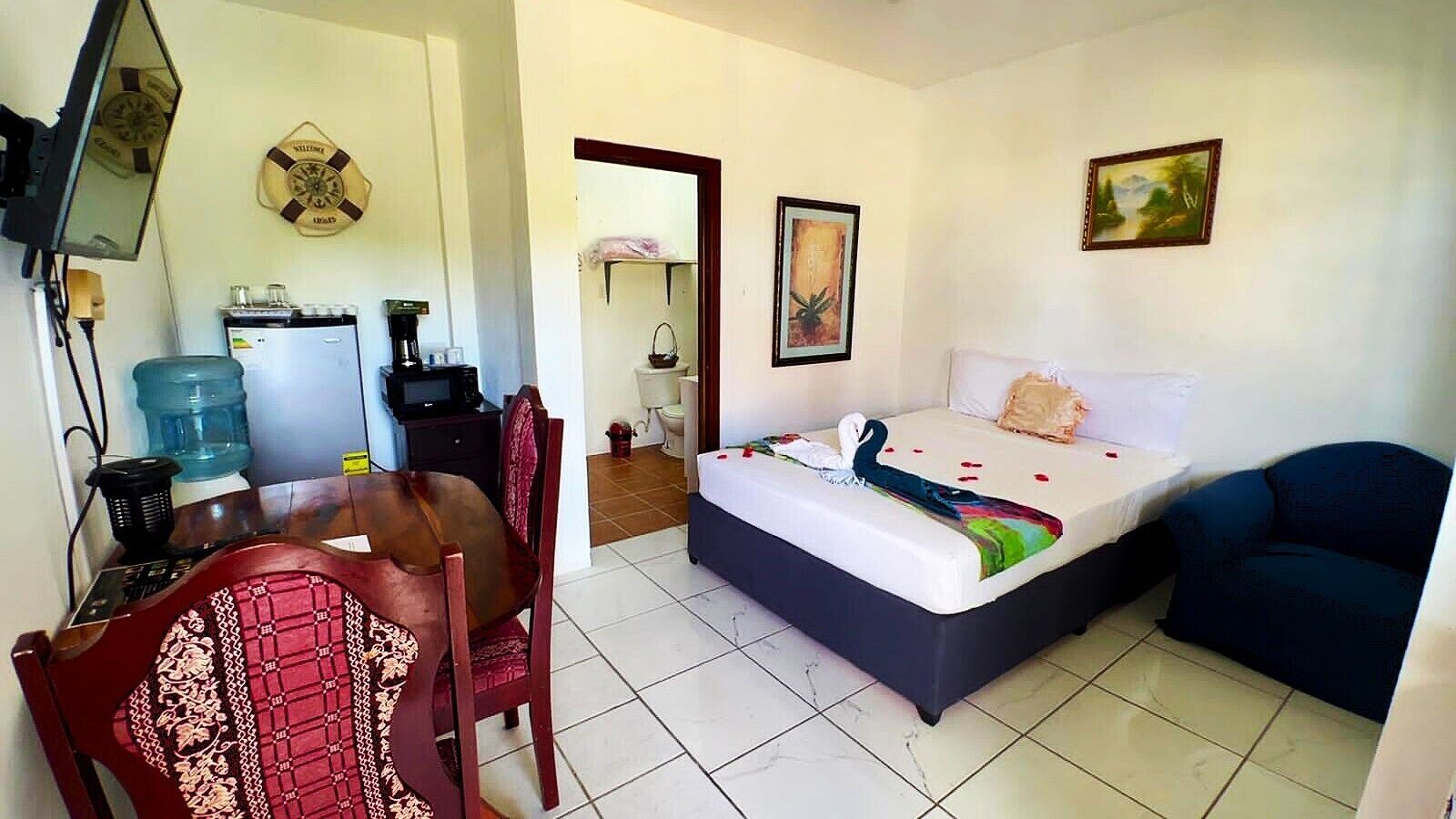 Habitación de hotel con cama, escritorio, silla, dispensador de agua, cafetera y un cuadro en la pared.