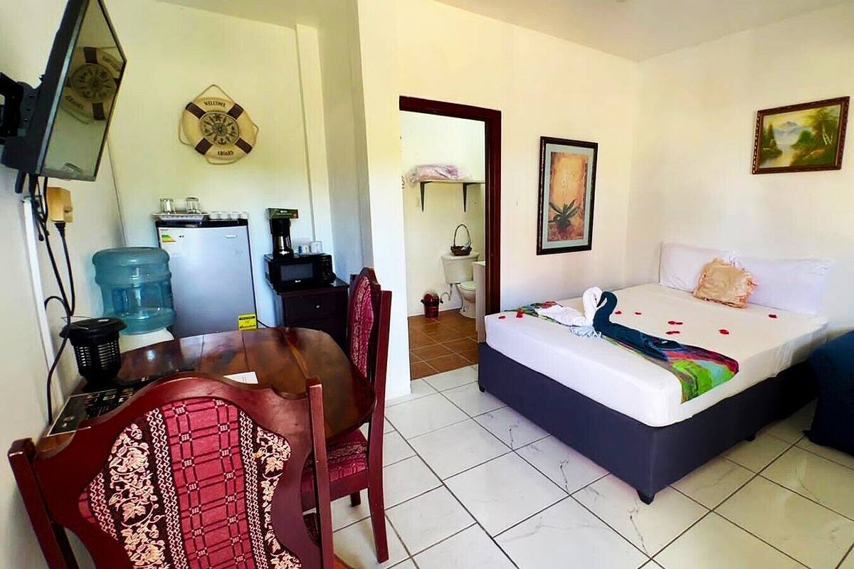 A hotel room with a bed, a desk, a chair, a water dispenser, a coffee machine, and a painting on the wall.