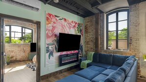 En stue med en blå sofa, et fladskærms-tv monteret på en blomstret væg, en murstensvæg og et stort vindue.