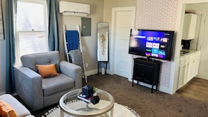 Een woonkamer met een grijze bank, een glazen salontafel, een tv op een dressoir, een ladder en een keukenruimte zichtbaar door een geopende deur.
