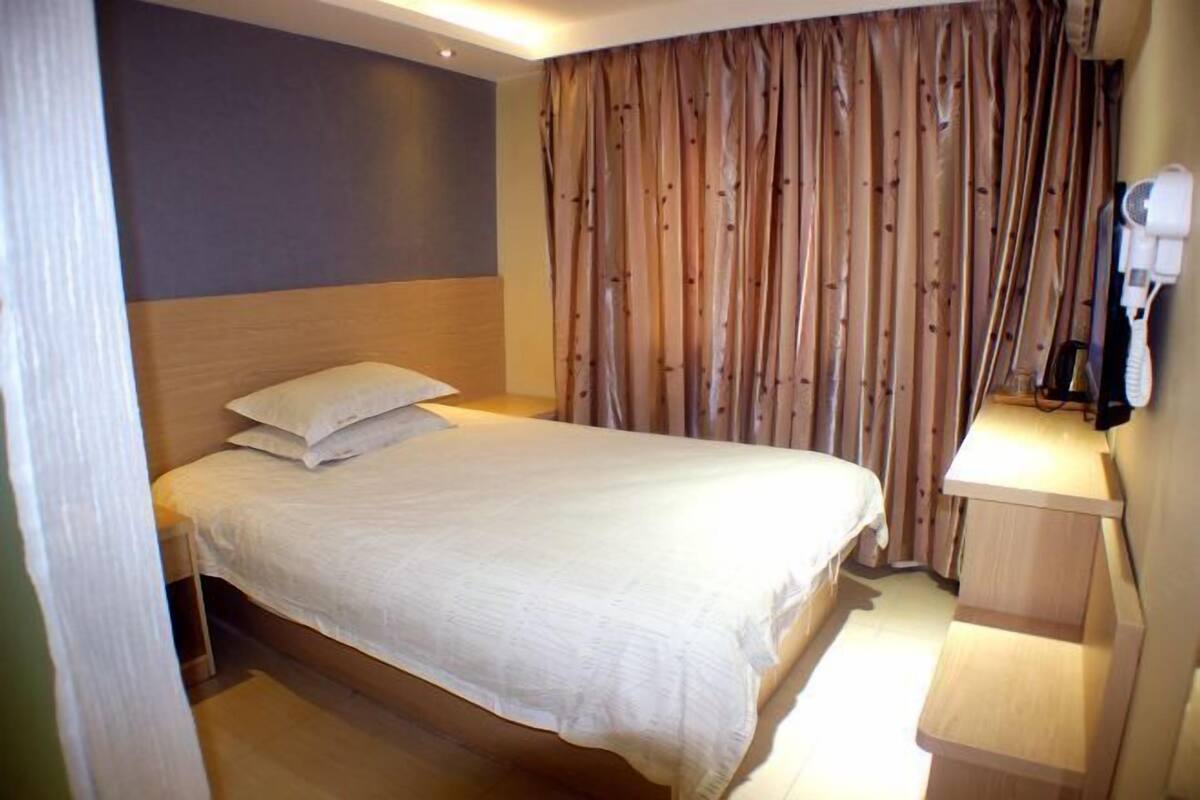Una cama individual con cabecera, una mesita de noche con lámpara y una cortina.