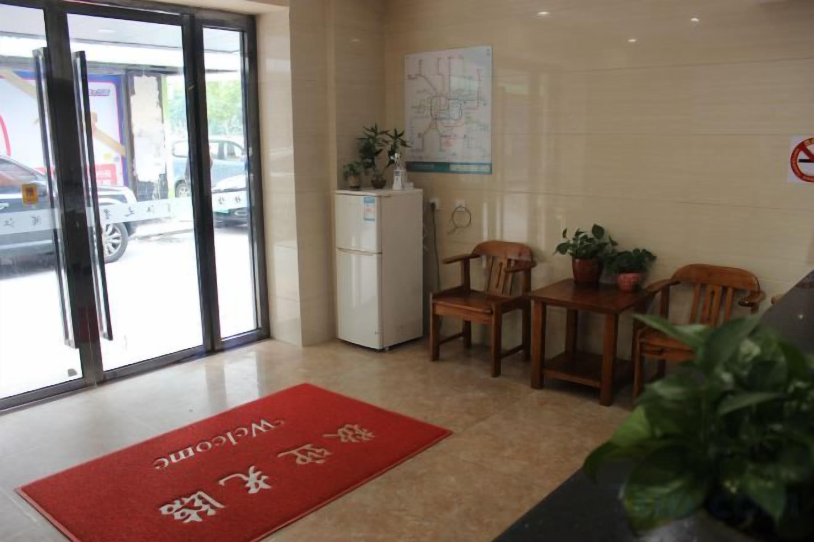 A hotel lobby with a reception desk, chairs, a refrigerator, and a red mat with text.