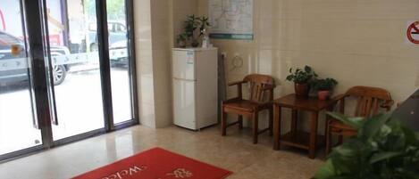 A hotel lobby with a reception desk, chairs, a refrigerator, and a red mat with text.