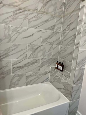 A bathroom with a marble tile wall, a white bathtub, and a shelf with toiletries.