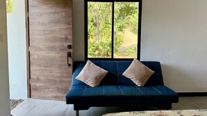 A room with a blue sofa, two pillows, a wooden door, and a window with a view of greenery. - Playa Mantas studio, AC, Parking (Quebrada Ganado)
