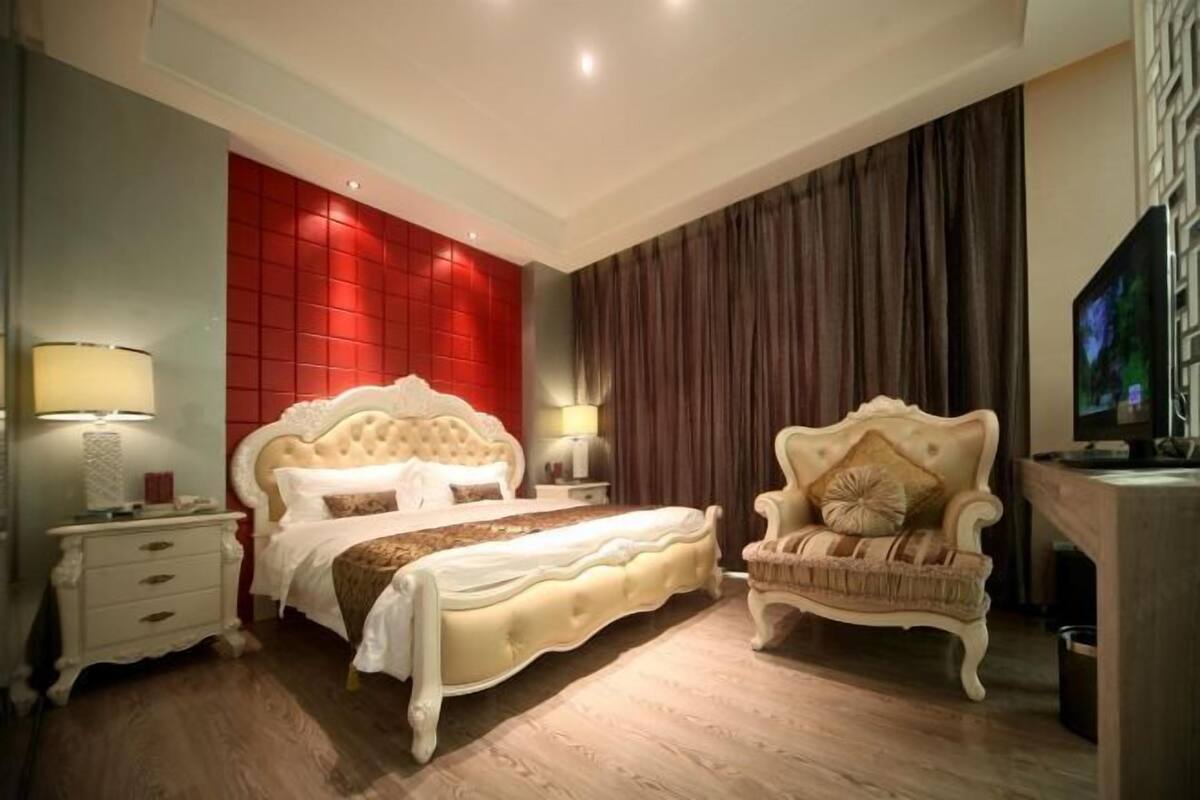 Ein Schlafzimmer mit einem großen Bett, einem Nachttisch, einem Sessel, einem Fernseher und einer roten Wand als Blickfang.