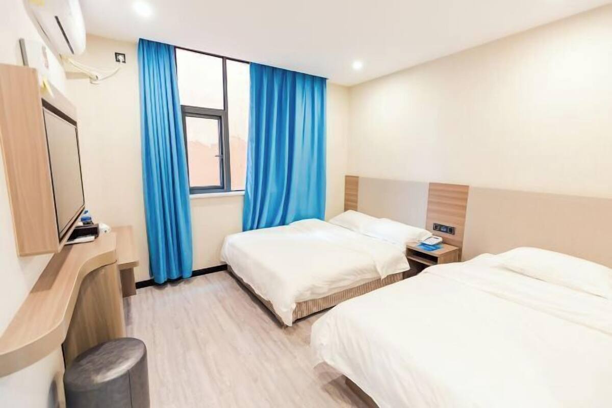 Habitación de hotel con dos camas, un escritorio con televisor de pantalla plana, una ventana con cortinas azules y un aire acondicionado instalado en la pared.