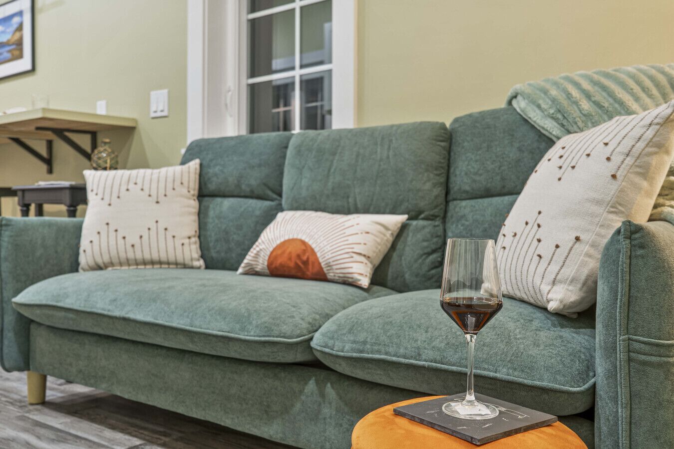 A green sofa with patterned pillows, a glass of red wine, and a small round table with a coaster.
