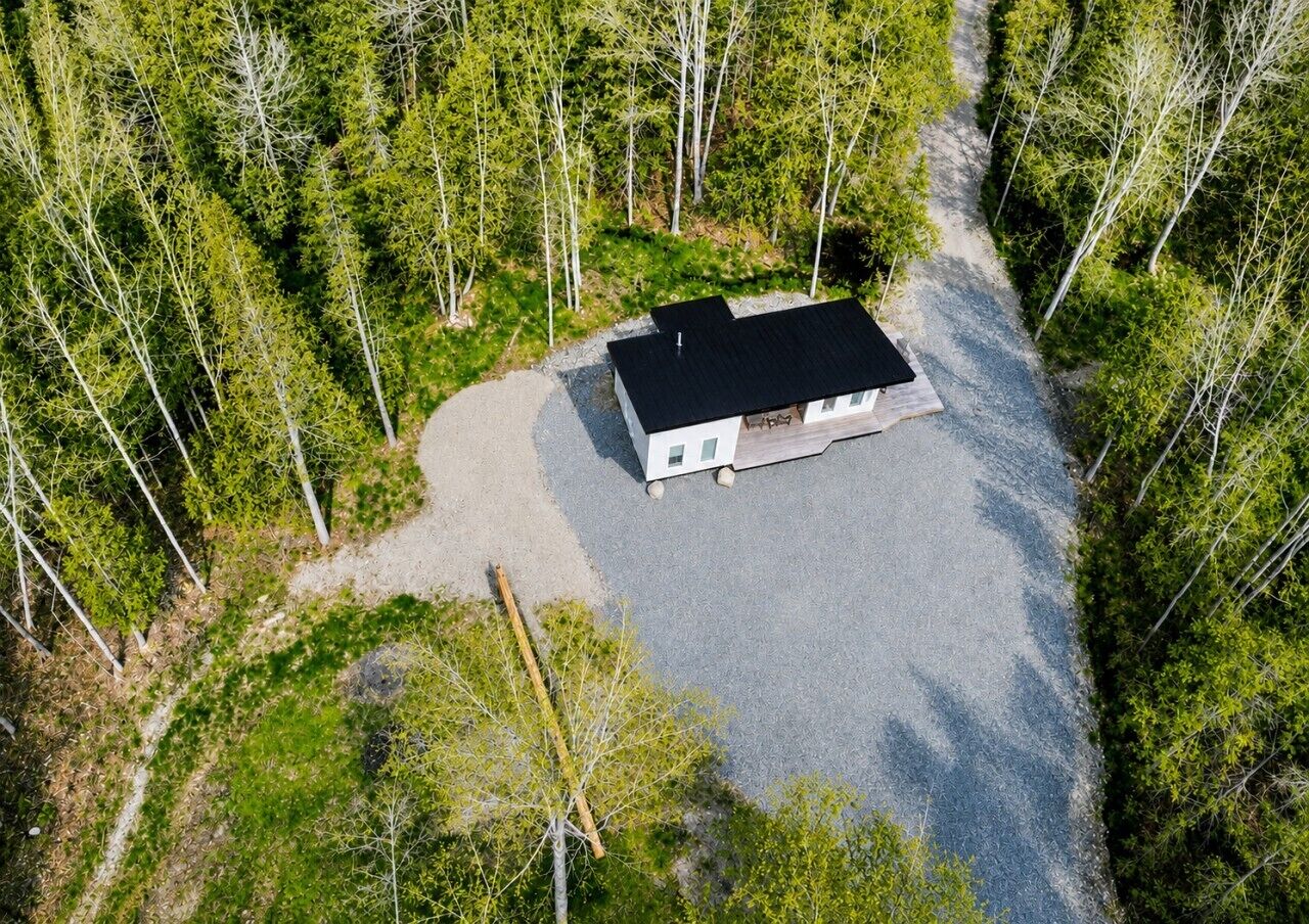 An aerial view of a single-story building with a black roof, surrounded by a gravel driveway and dense forest.