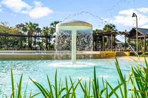 A fountain with water jets in a pool area, surrounded by palm trees and a gazebo.