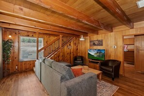En ombonad träinredning med en trappa, en grå soffa, en svart läderfåtölj, en tv och en tavla på väggen.