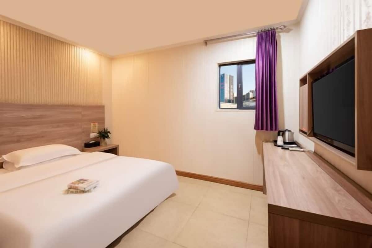 A hotel room with a bed, a TV, a window with curtains, and a wooden headboard.