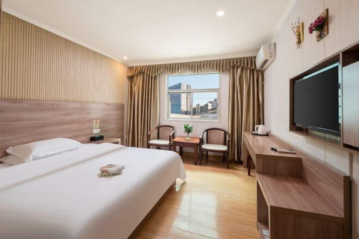 A hotel room with a large bed, a desk with a TV, a small table with chairs, and a window with curtains.
