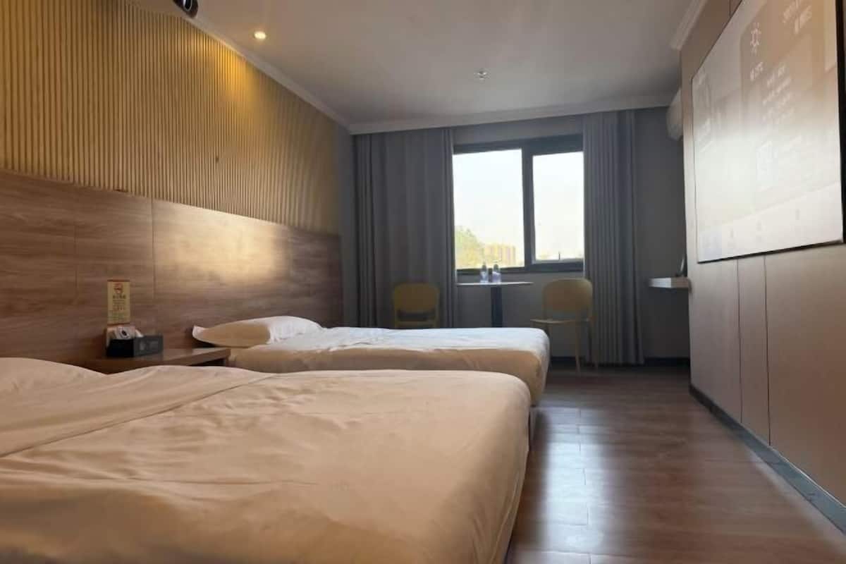 A hotel room with two beds, a wooden headboard, a small table with chairs, and a large window with curtains.