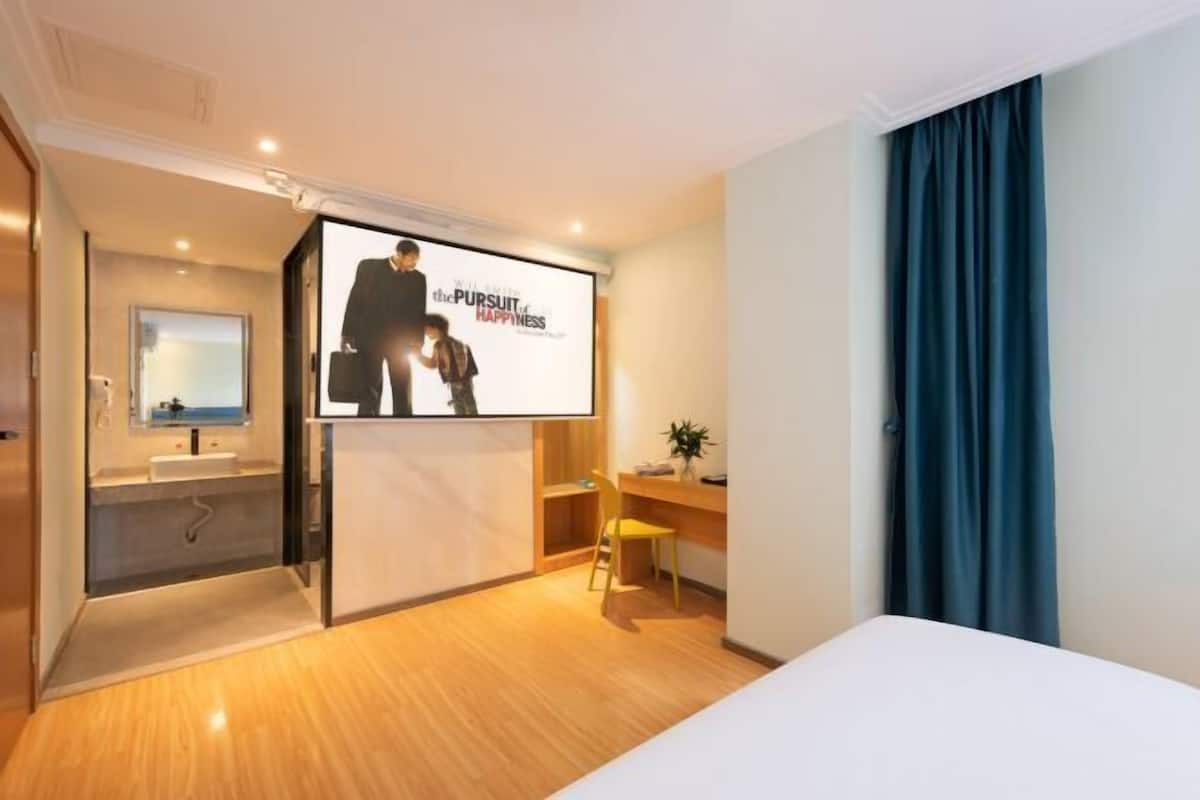 A modern hotel room with a large bed, a desk, a chair, a small sink, and a large window with curtains.