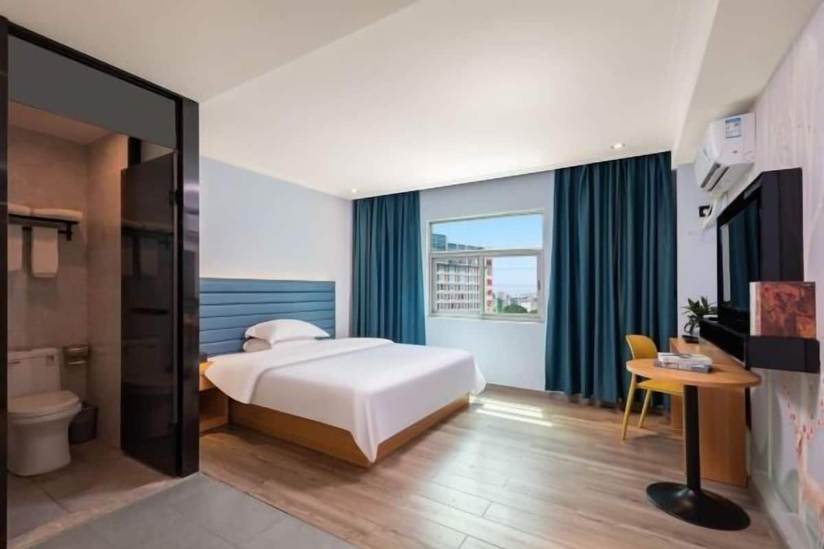 A hotel room with a bed, a desk, a chair, a window with curtains, and an air conditioning unit.