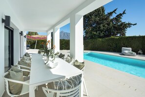 Un espace de restauration extérieur moderne, doté d’une piscine, de mobilier blanc et offrant une vue sur les montagnes.