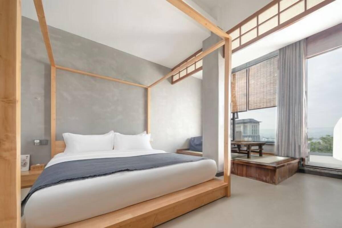 A modern bedroom with a wooden four-poster bed, a small table, and a view of the city.