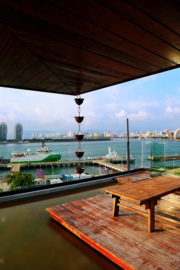 Una terraza de madera con un banco, una lámpara colgante y vistas a un puerto deportivo con barcos y el perfil de una ciudad al fondo.