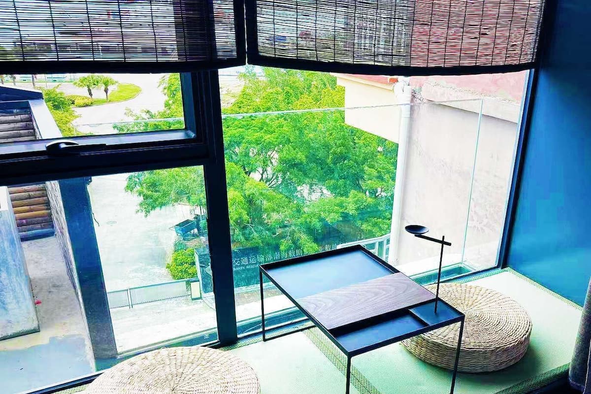 Ein Zimmer mit Blick auf Grün hinter einem Fenster, ein kleiner Tisch und zwei runde, gewebte Poufs.