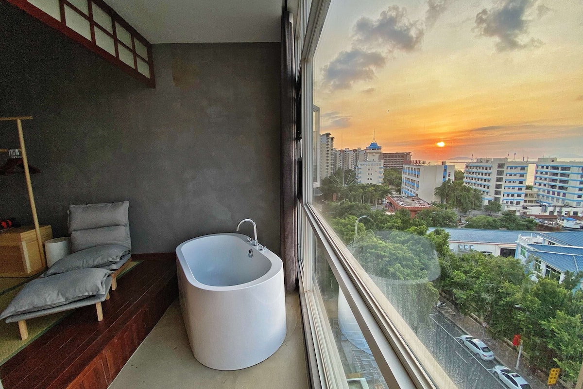 Ein Badezimmer mit einer Badewanne, einer Toilette und einem Fenster mit Blick auf eine Stadtlandschaft bei Sonnenuntergang.