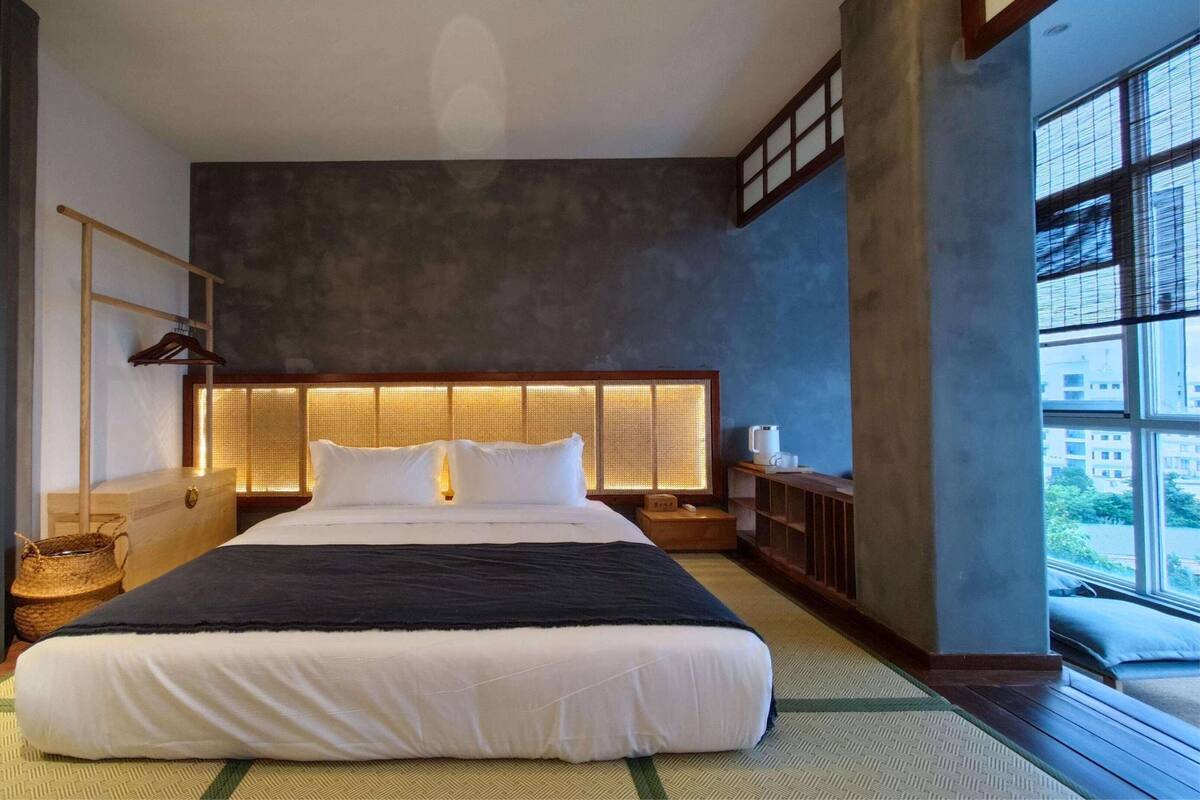 Ein Hotelzimmer mit einem großen Bett, einem hölzernen Kopfteil, einem kleinen Nachttisch mit Lampe und Blick auf die Stadt durch große Fenster.