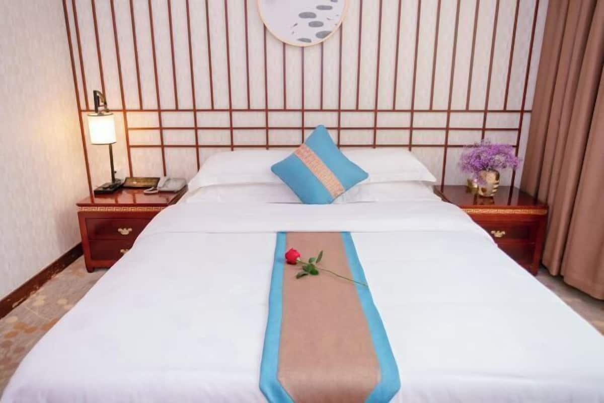 Habitación de hotel con una cama, una mesita de noche con lámpara, un florero con flores y una pared con líneas verticales.