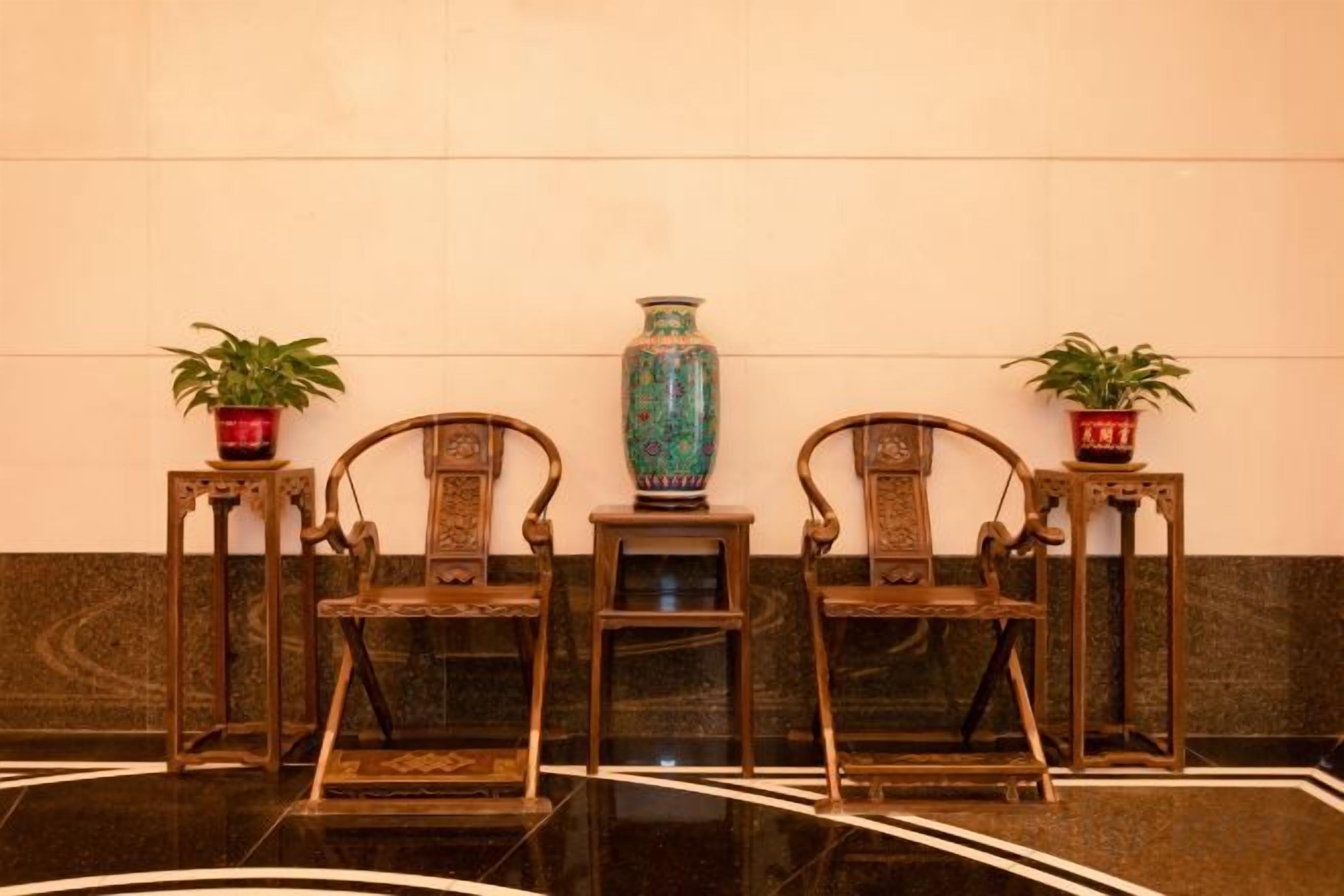 A lobby with three wooden chairs, a large blue vase, and two potted plants on side tables.