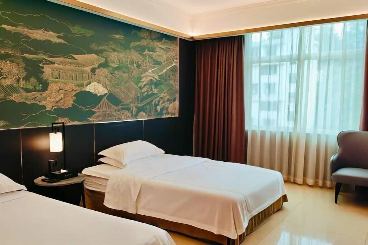 Habitación de hotel con dos camas, un mural grande, un escritorio con lámpara, una silla y un ventanal con cortinas.
