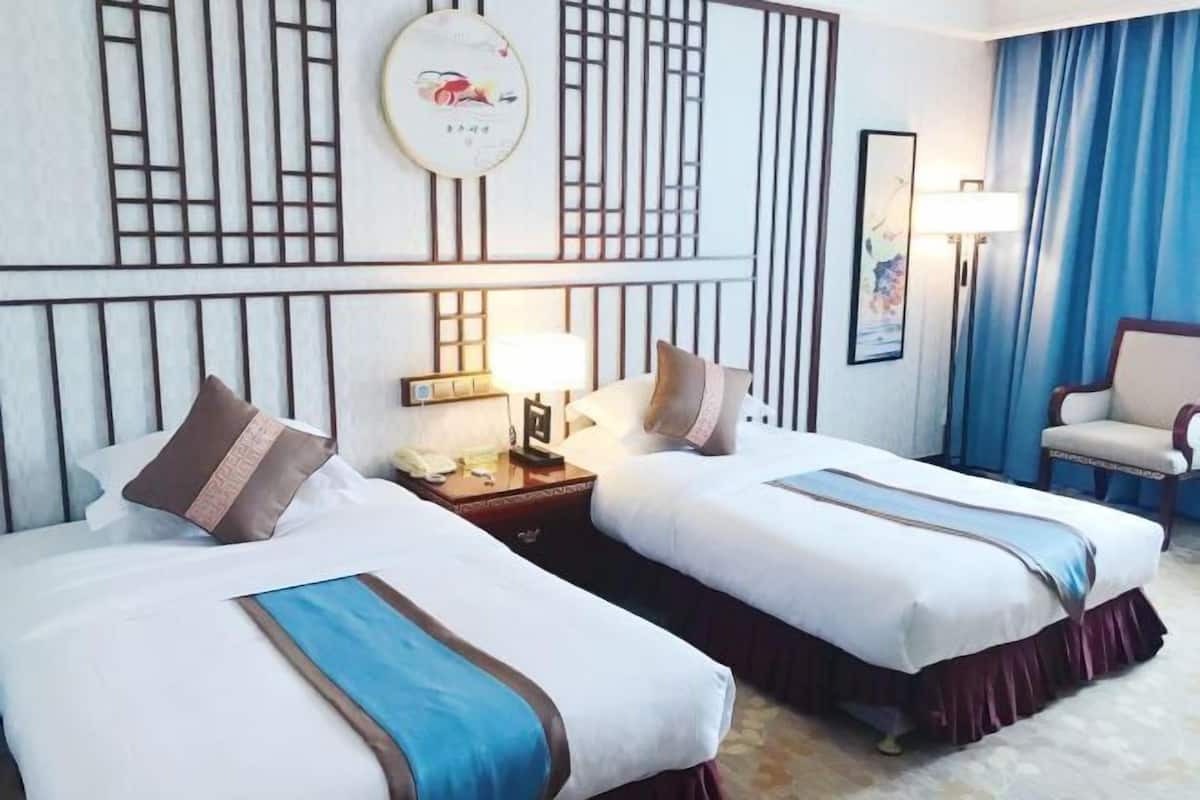 Habitación de hotel con dos camas, una mesita de noche con lámpara, una silla y un panel decorativo en la pared.