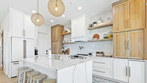 A modern kitchen with white cabinetry, a large island with stools, wooden accents, and pendant lighting. - Luxury 5 BR Beach House - Lagoon View Heated Pool (Gulf Shores)