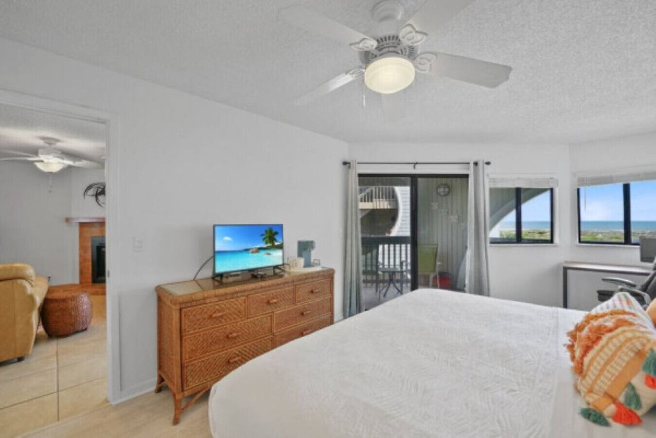 A bedroom with a bed, a television on a wooden dresser, a ceiling fan, and a balcony with a view of the ocean.