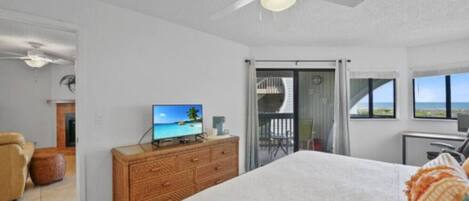 A bedroom with a bed, a television on a wooden dresser, a ceiling fan, and a balcony with a view of the ocean.