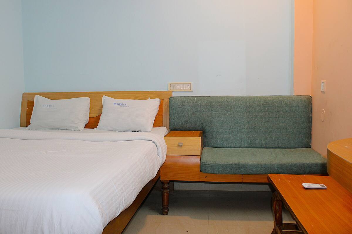 Una cama bien tendida con sábanas blancas, un sillón tapizado en verde y una mesita de noche de madera con un control remoto.