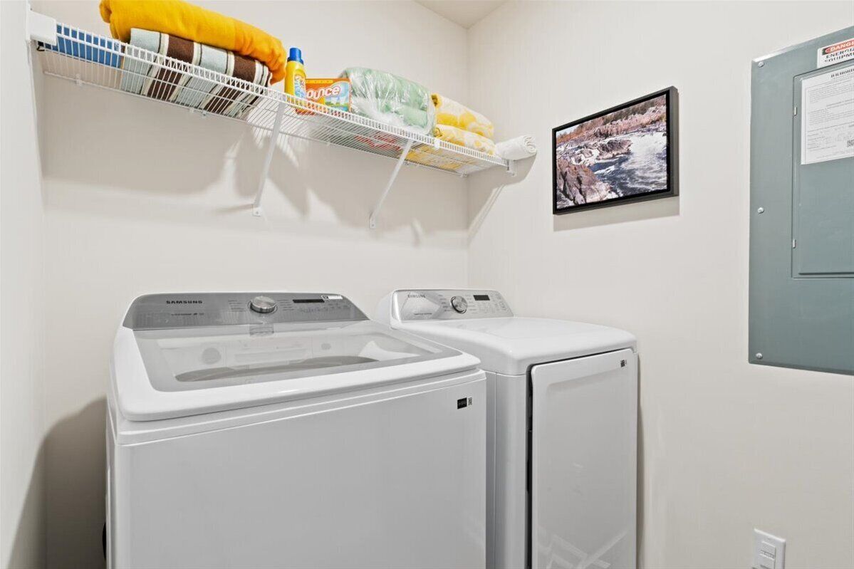 A laundry room with two washers and dryers, a shelf with laundry supplies, and a picture on the wall.