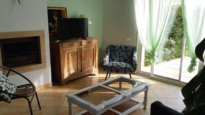A living room with a fireplace, a television on a wooden cabinet, a patterned armchair, and a glass coffee table. - Gite Camélias 700 m from the sea and the beach, 3 stars, Portsall, GR34 (Ploudalmézeau)
