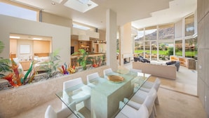 A spacious, modern interior with a glass dining table, white chairs, and a view of a landscaped outdoor area.