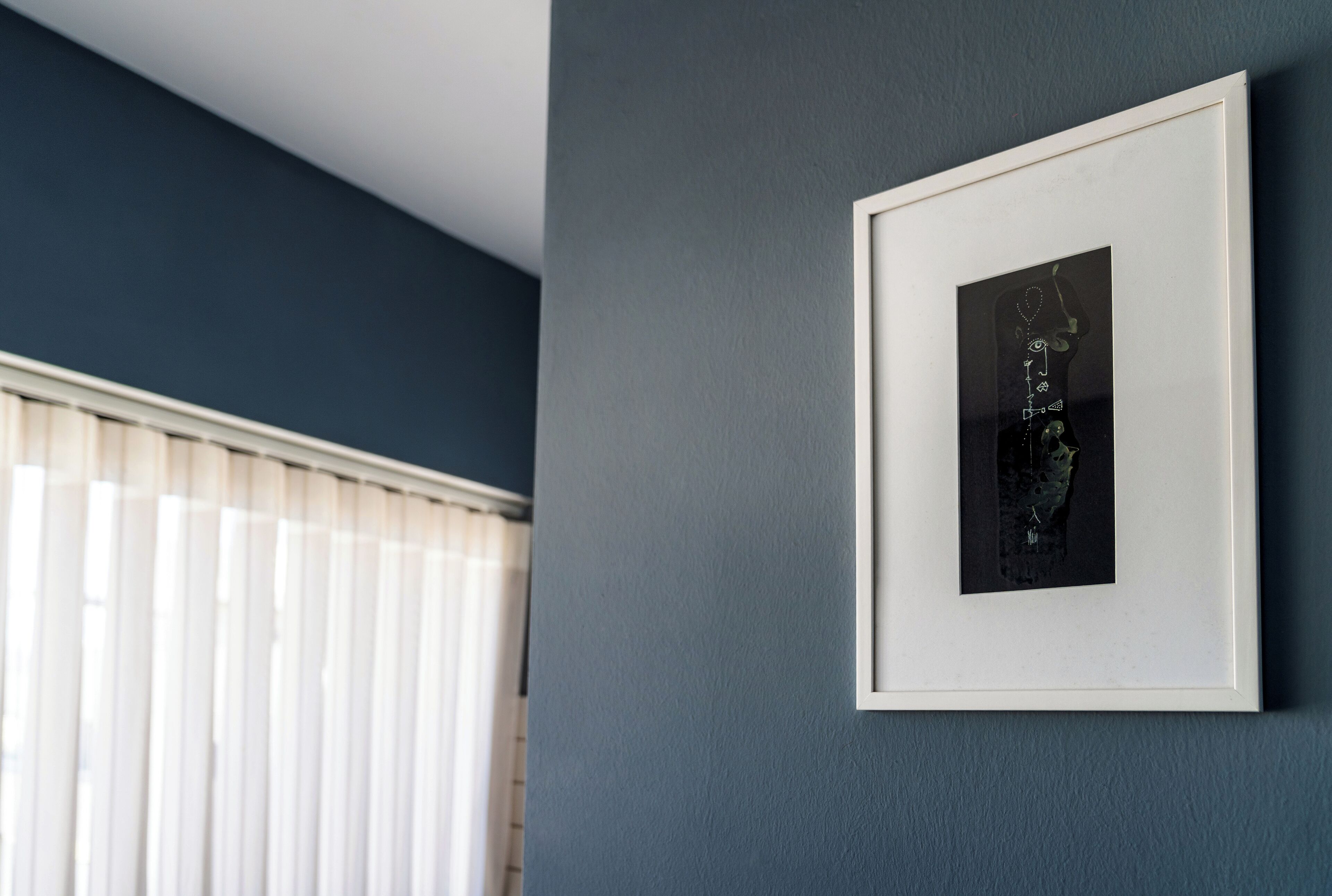 Une œuvre d’art encadrée accrochée au mur, une fenêtre avec des stores et un mur bleu foncé.