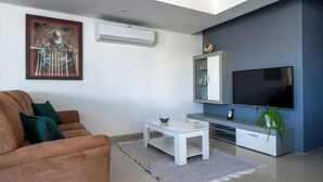 A modern living room with a brown sofa, a white coffee table, a flat-screen TV, a small cabinet, and a potted plant. - Bellavista Bayview Penthouse Terrace (Marsaskala)