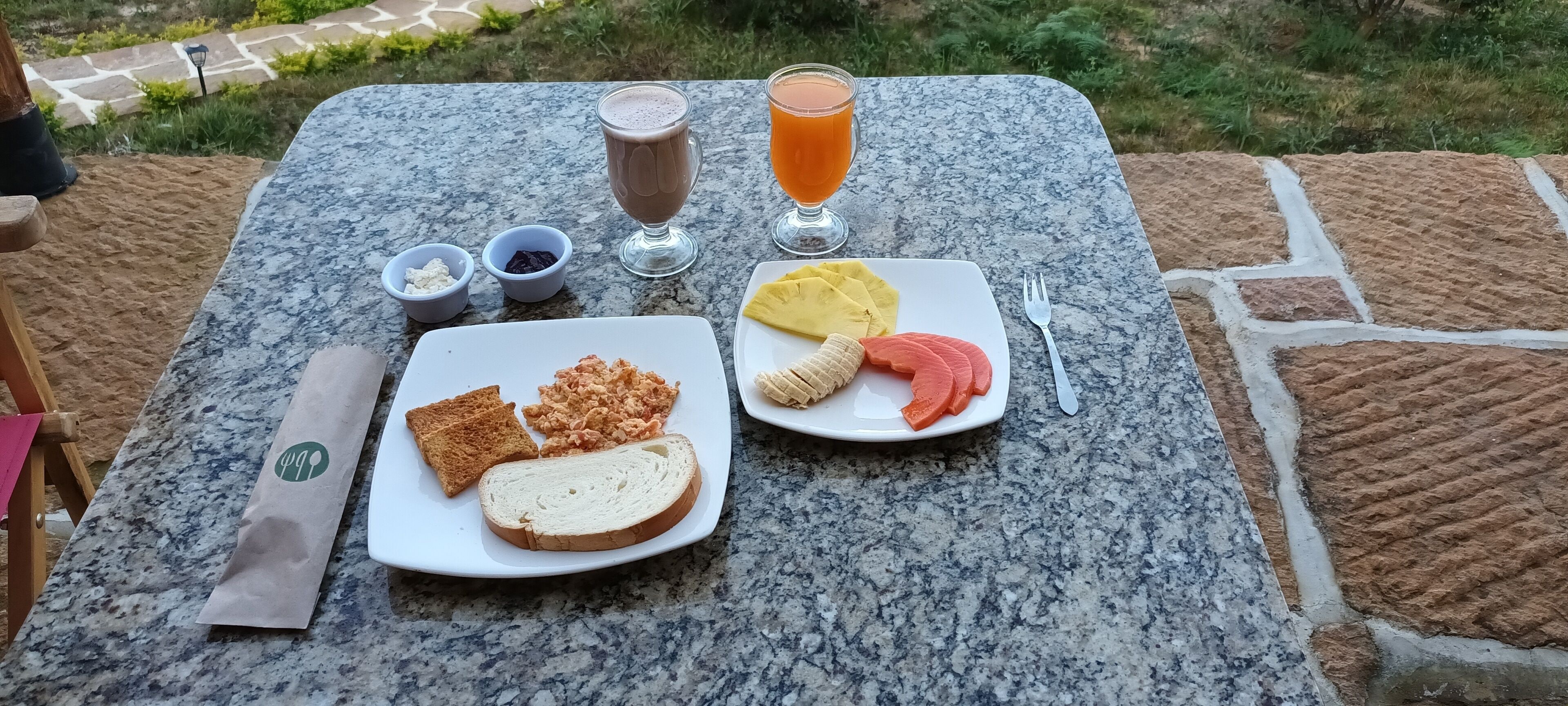 Un montaje de comida en una mesa exterior con superficie de baldosas de piedra, que incluye tostadas, un plato con fruta y una bebida.