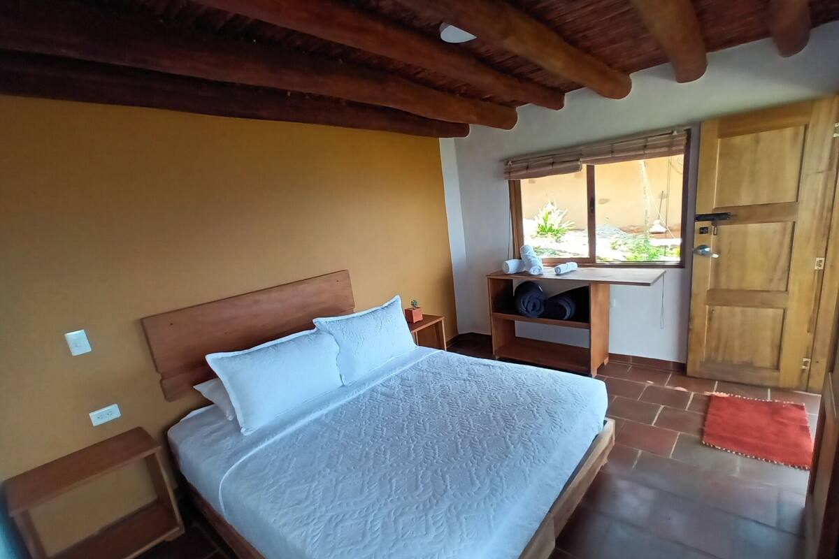 A bedroom with a wooden headboard, a bed with white linens, a wooden nightstand, a shelf with rolled towels, a window with a view, and a wooden door.