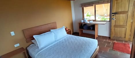 A bedroom with a wooden headboard, a bed with white linens, a wooden nightstand, a shelf with rolled towels, a window with a view, and a wooden door.