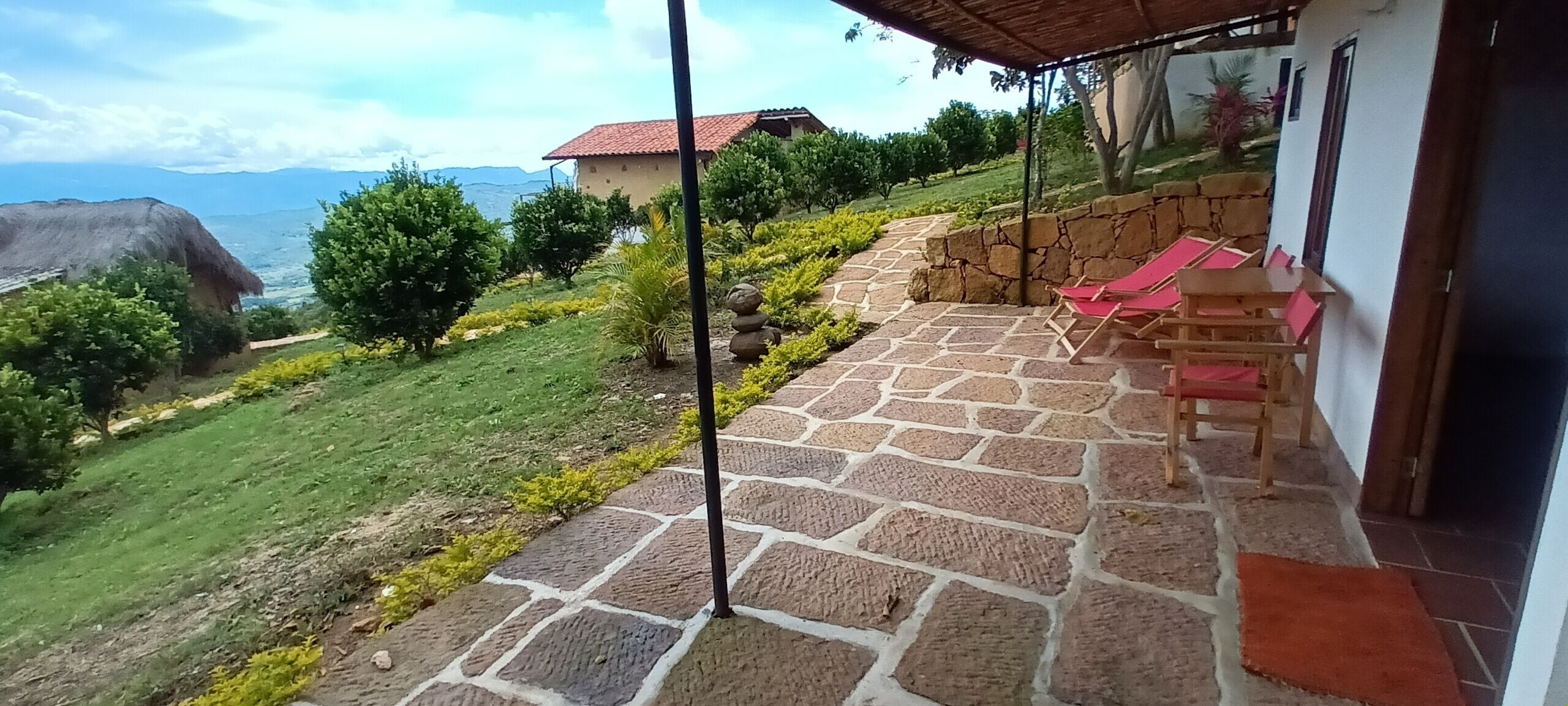 Un patio empedrado con sillas rojas, un muro de piedra y vistas a una colina con árboles y un edificio distante.